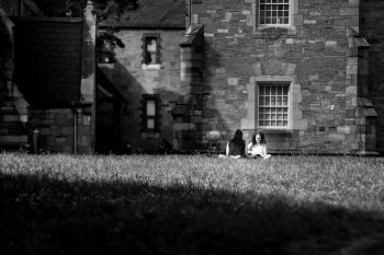 Two people sit on grass near an old stone building