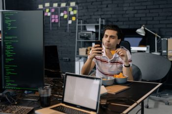 A programmer checks his smartphone while eating snacks at a modern tech office desk